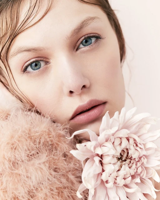 Close-up portrait of a fair-skinned young woman with blue eyes and wet-look hair holding a large pink dahlia flower against a pale pink background.
