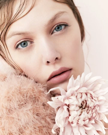 Close-up portrait of a fair-skinned young woman with blue eyes and wet-look hair holding a large pink dahlia flower against a pale pink background.