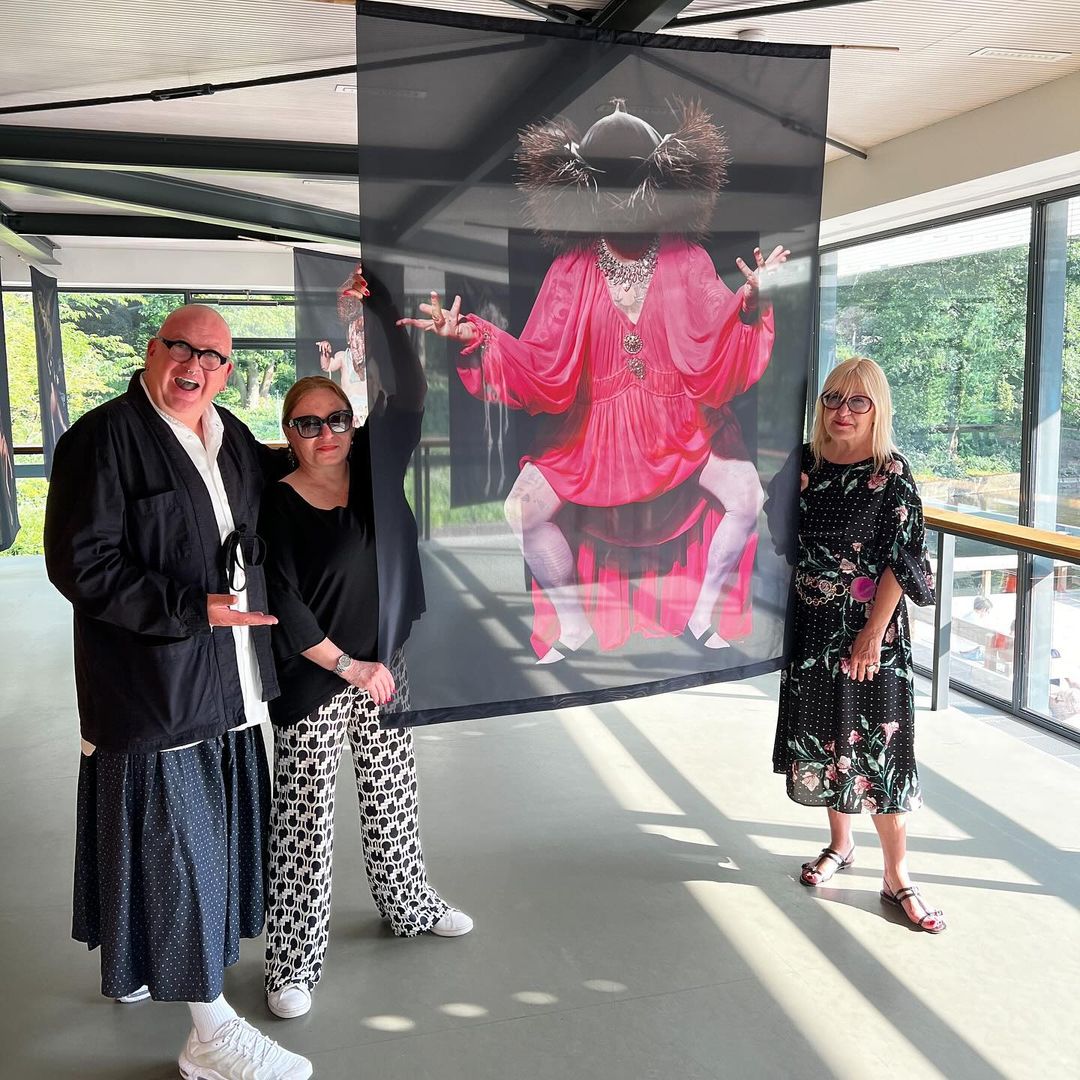 Three adults pose next to an indoor art installation of a figure in a red dress, inside a building with a window to a green background.