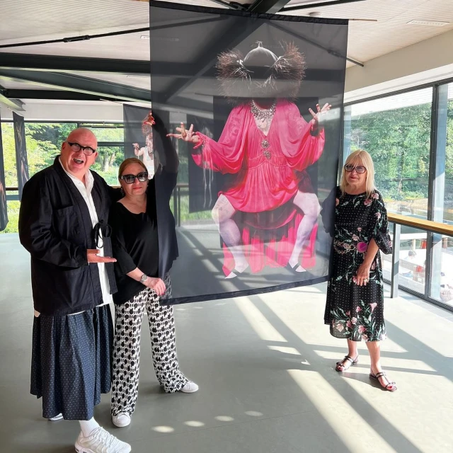 Three adults pose next to an indoor art installation of a figure in a red dress, inside a building with a window to a green background.