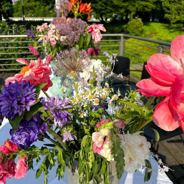 Outdoor display of colorful peonies, daisies, and allium flowers arranged in a metal vase on a sunny day.