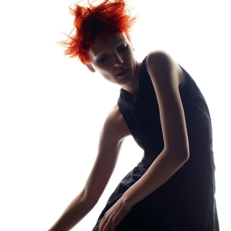 Model with vibrant spiky red hair wearing a dark sleeveless dress against a bright white background