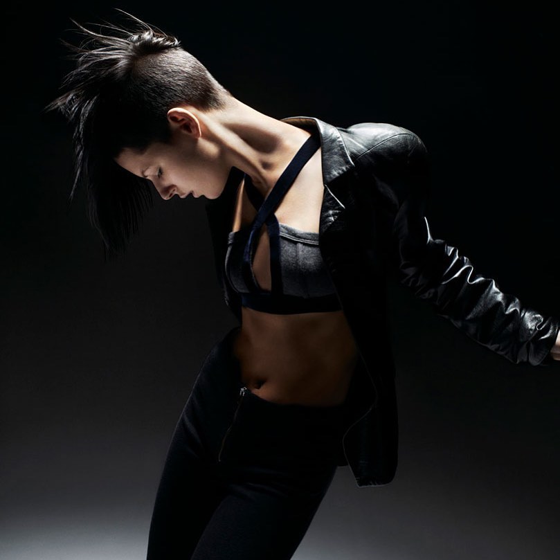 Woman in edgy fashion poses in photo studio. She wears a black leather jacket and matching top and pants in a dramatic pose.