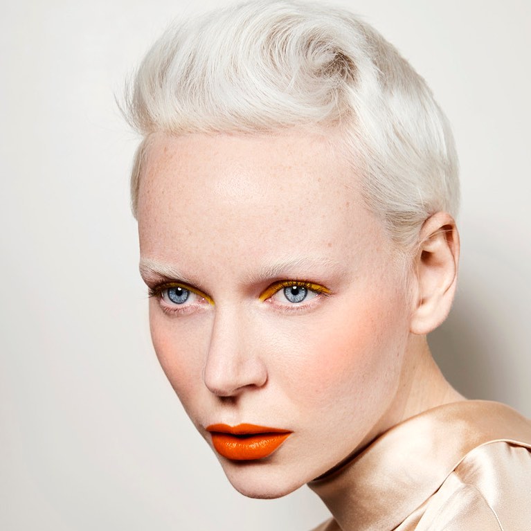 Close-up portrait of a fair-skinned adult female with short platinum-blonde hair, bright blue eyes, orange lipstick and yellow eyeliner.