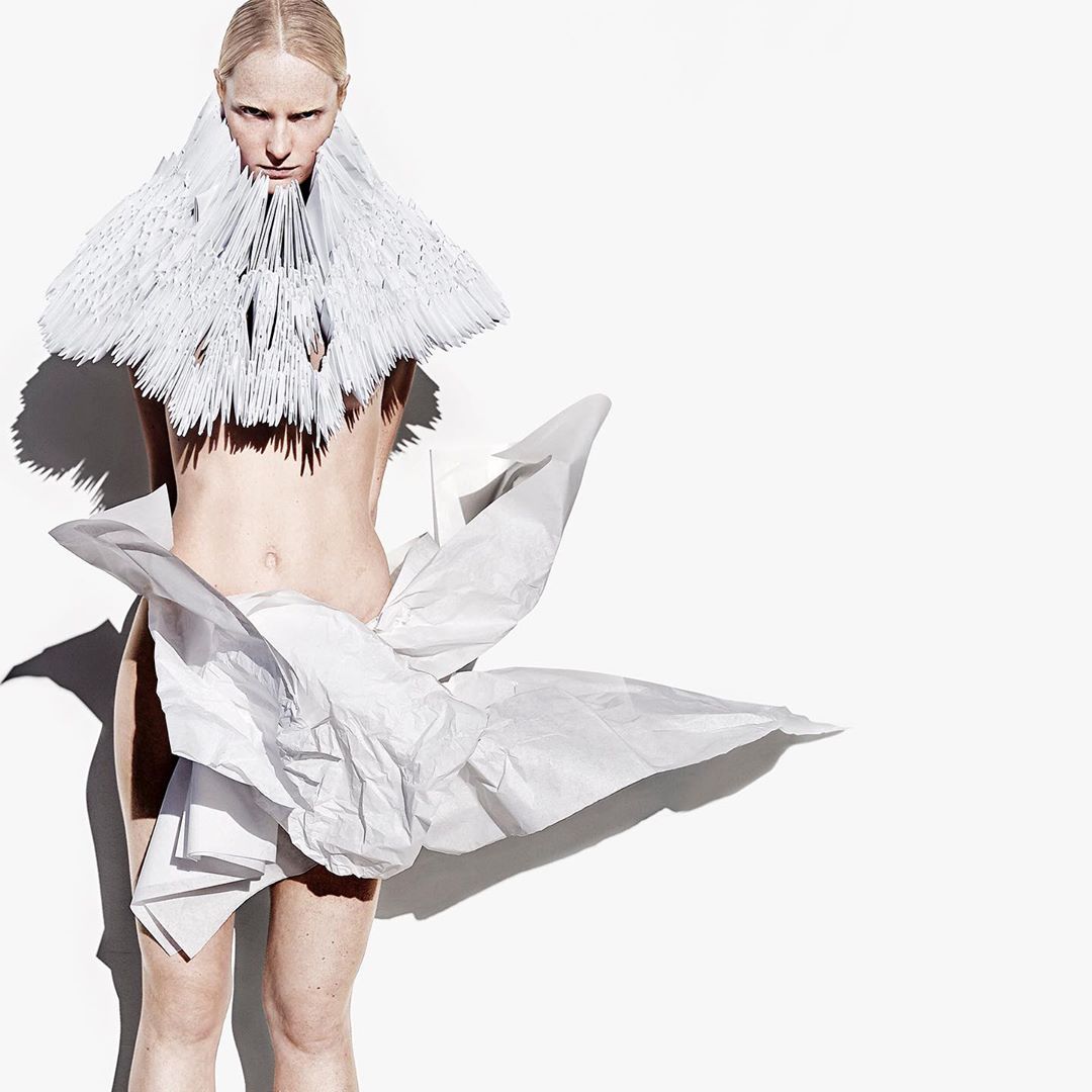 Artistic fashion photo of a fair-skinned person wearing an elaborate handmade paper dress and collar against a bright white backdrop.