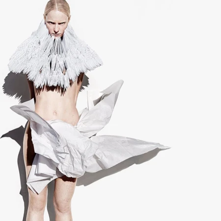 Artistic fashion photo of a fair-skinned person wearing an elaborate handmade paper dress and collar against a bright white backdrop.