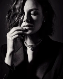 Close-up portrait of a woman with dark wavy hair wearing a diamond necklace and dark blazer, captured in dramatic low-key lighting.
