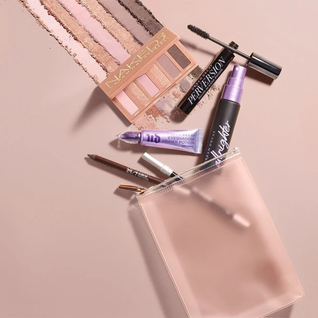 Makeup flat lay featuring Urban Decay Naked eyeshadow palette, Perversion mascara, All Nighter setting spray, and a clear makeup bag.