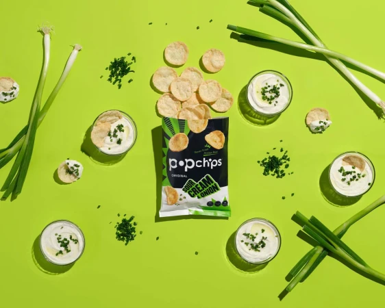 Overhead shot of Popchips Sour Cream & Onion chips with chives, scallions, and dip on a bright green background, food photography.