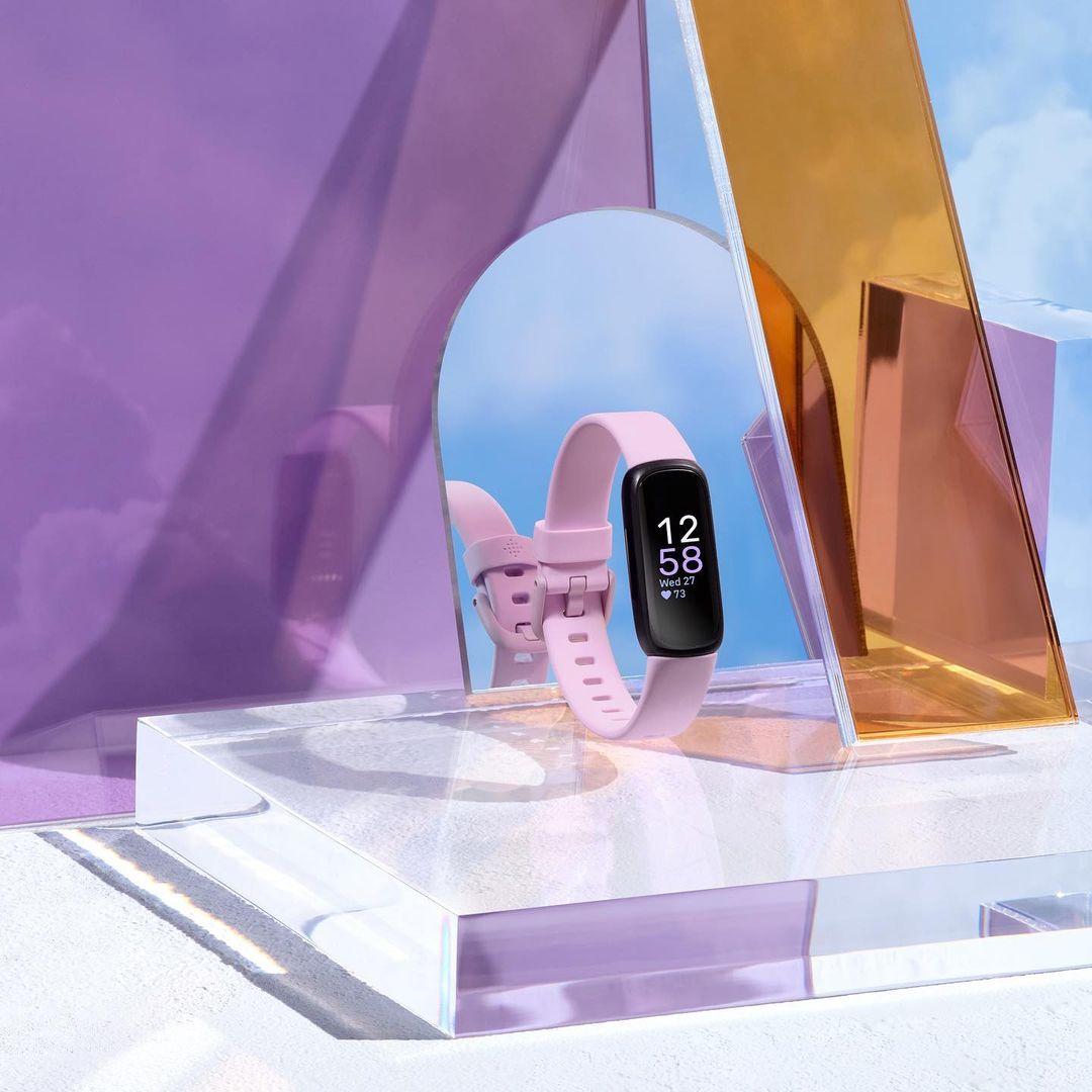 Close-up studio shot of a pink fitness tracker on a clear acrylic platform, reflecting against colorful geometric mirrors.
