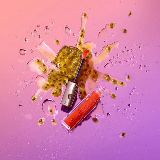 Urban Decay orange lip gloss laying on a glossy purple surface with passion fruit and water droplets, beauty product shot.