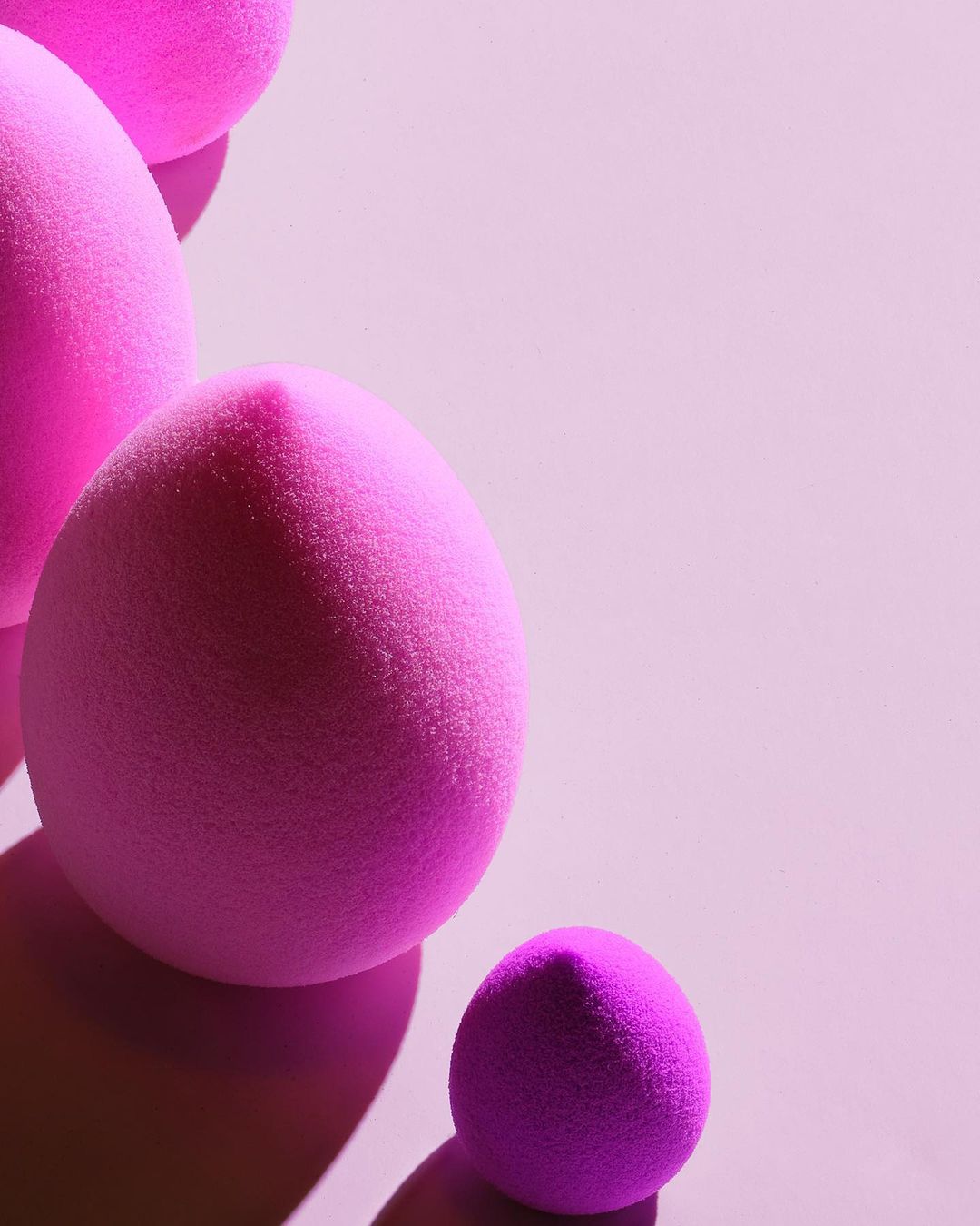 Close-up of pink makeup sponges in varying sizes, arranged on a soft pink background with shadows and space for text or graphics.