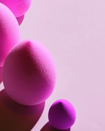 Close-up of pink makeup sponges in varying sizes, arranged on a soft pink background with shadows and space for text or graphics.