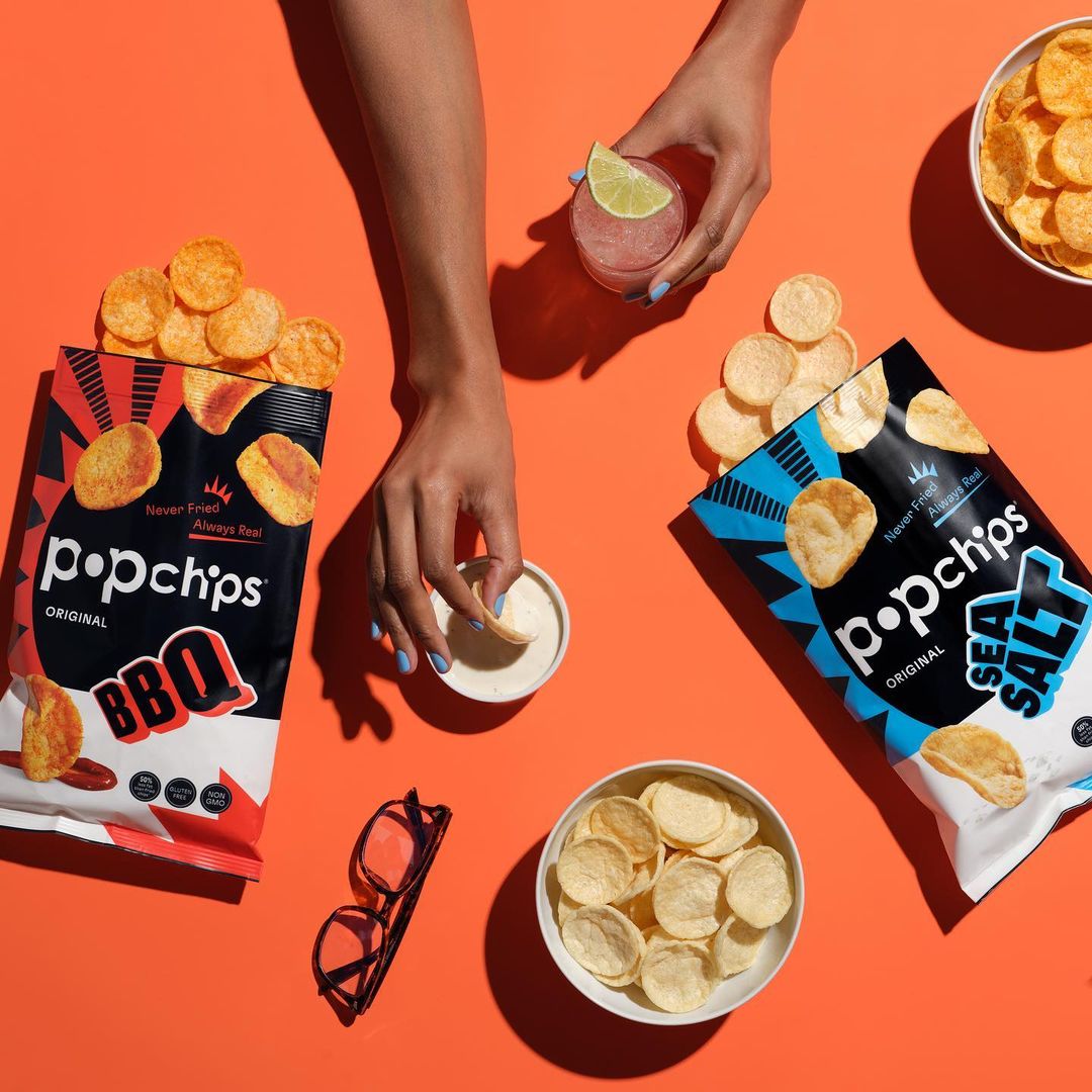 Overhead shot of Popchips bags, a clear cocktail, and small bowls of chips and dip on a vibrant orange surface, showcasing snack presentation.