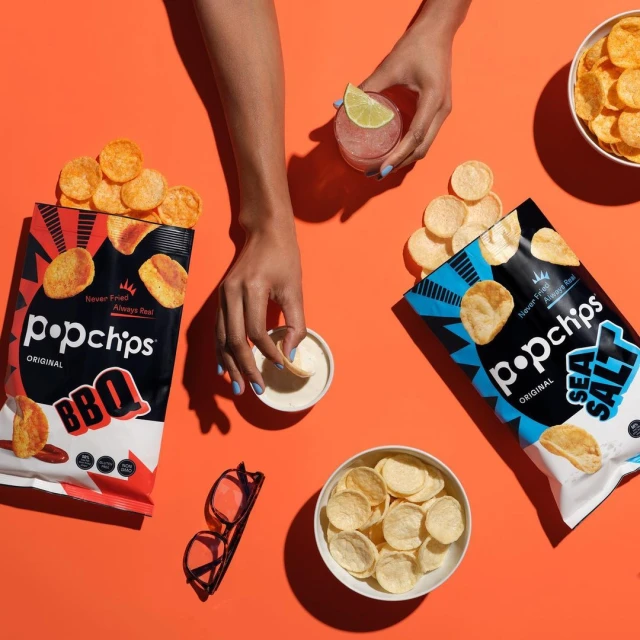 Overhead shot of Popchips bags, a clear cocktail, and small bowls of chips and dip on a vibrant orange surface, showcasing snack presentation.
