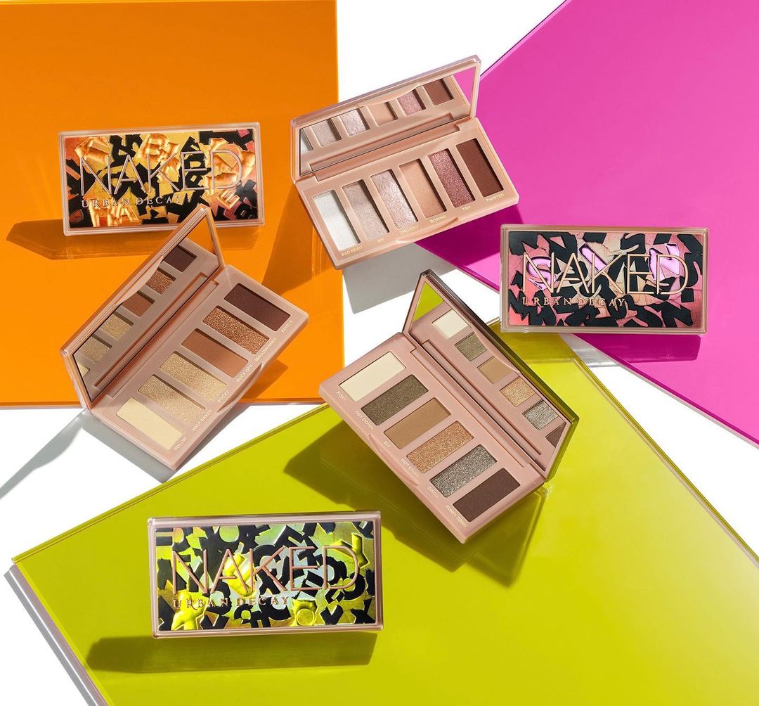 Multiple eyeshadow palettes from Urban Decay's Naked collection are arranged on vibrant orange, pink, and green surfaces. Beauty product photography.