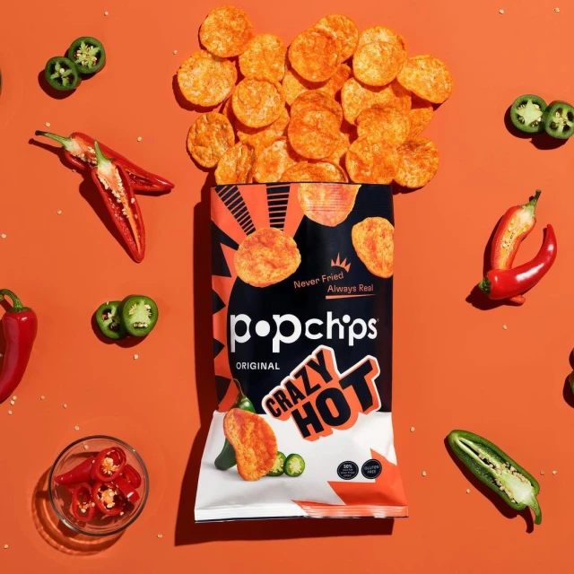 Overhead shot of Popchips Crazy Hot chips bag with chips, jalapeños, and red chili peppers against an orange background.