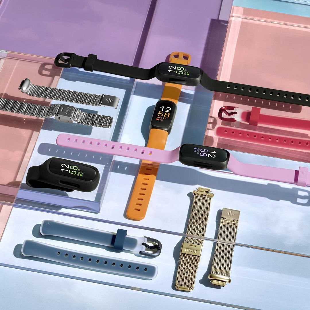 Multiple Fitbit fitness trackers and bands in various colors and styles are arranged on a translucent surface for a product showcase.
