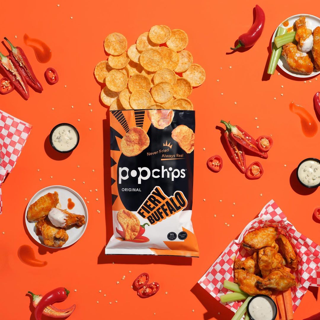 Popchips Fiery Buffalo chips bag surrounded by chips, hot wings, and sliced red peppers on an orange background. Food and beverage.