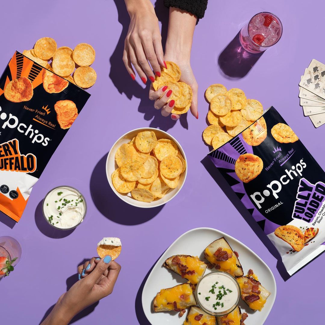 Overhead shot of Popchips snacks and dips on a purple surface, with hands holding and serving the chips, creating an inviting food flat lay.
