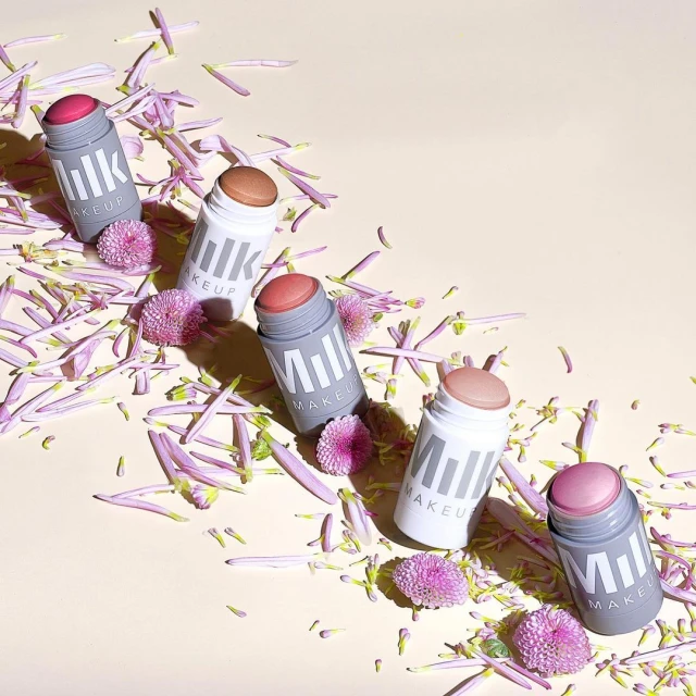 Five Milk MAKEUP sticks in assorted colors are arranged amidst purple flower petals against a light peach background, product photography.