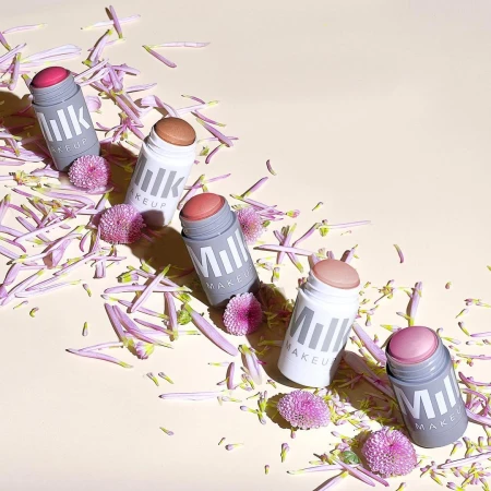 Five Milk MAKEUP sticks in assorted colors are arranged amidst purple flower petals against a light peach background, product photography.