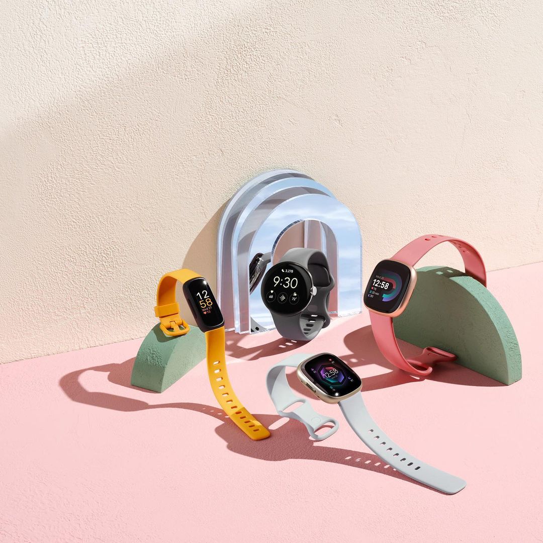 Five colorful smartwatches with unique bands are arranged on geometric stands against a split pink and beige textured background.