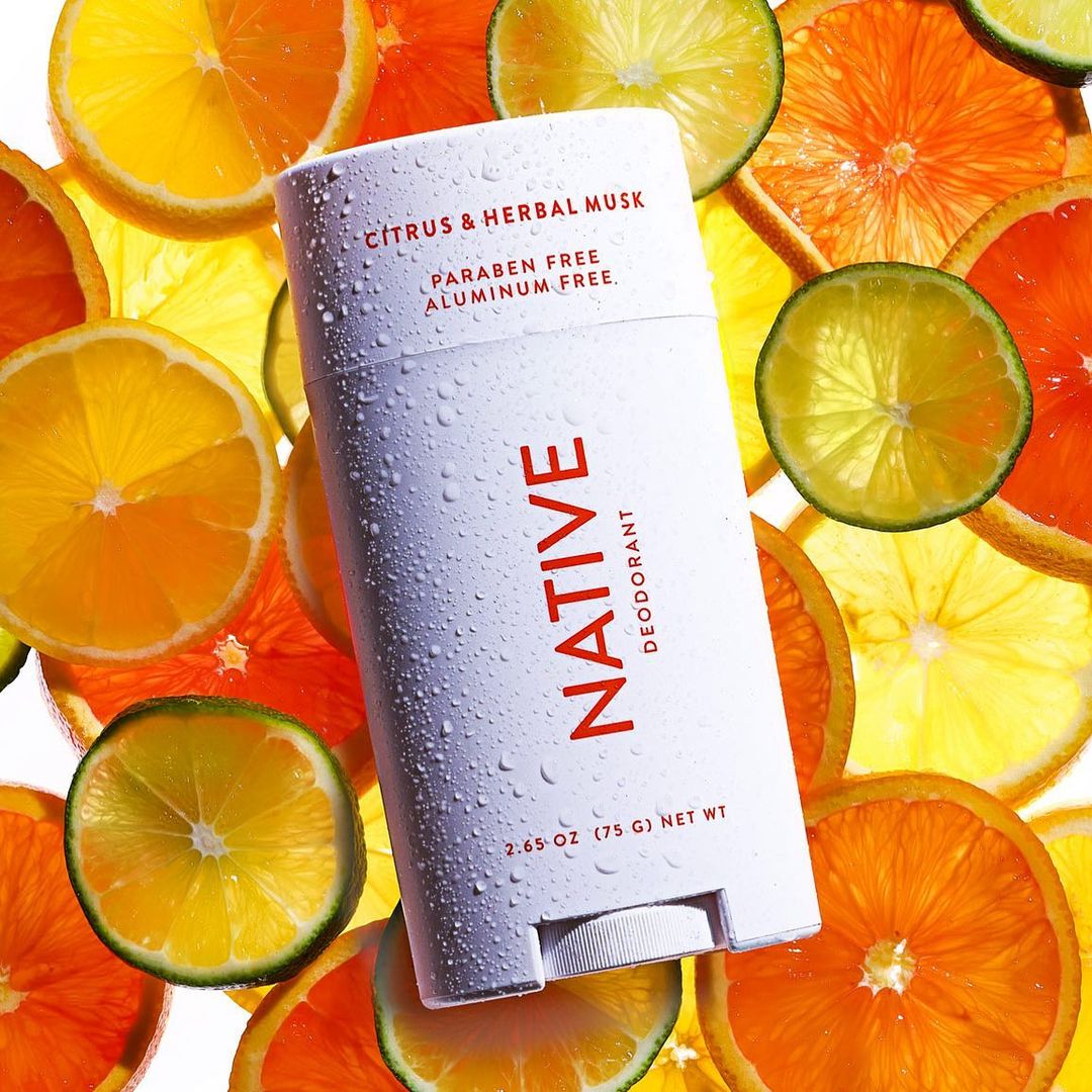 Close-up of Native deodorant stick amidst sliced citrus fruits. White packaging with red lettering, promoting natural personal care.