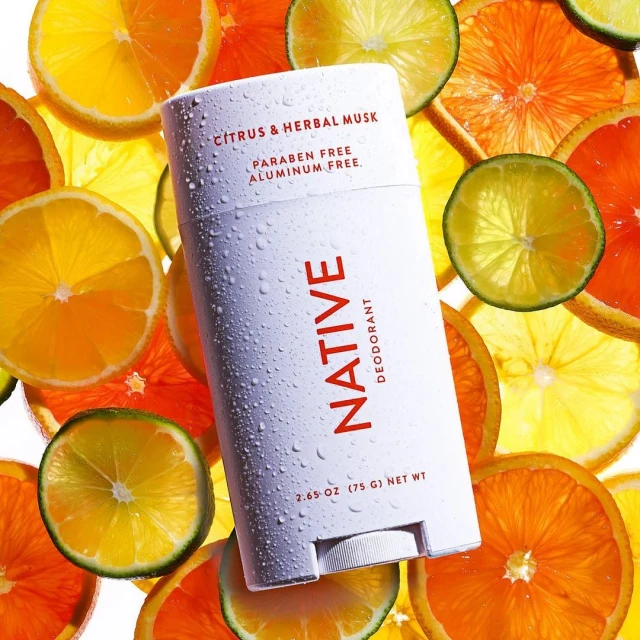 Close-up of Native deodorant stick amidst sliced citrus fruits. White packaging with red lettering, promoting natural personal care.