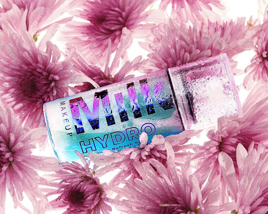 Milk Makeup Hydro product with water droplets, nestled amongst a bed of pink chrysanthemum flowers, close-up beauty shot on white.