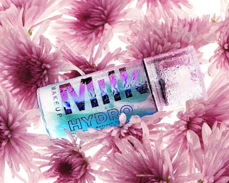 Milk Makeup Hydro product with water droplets, nestled amongst a bed of pink chrysanthemum flowers, close-up beauty shot on white.