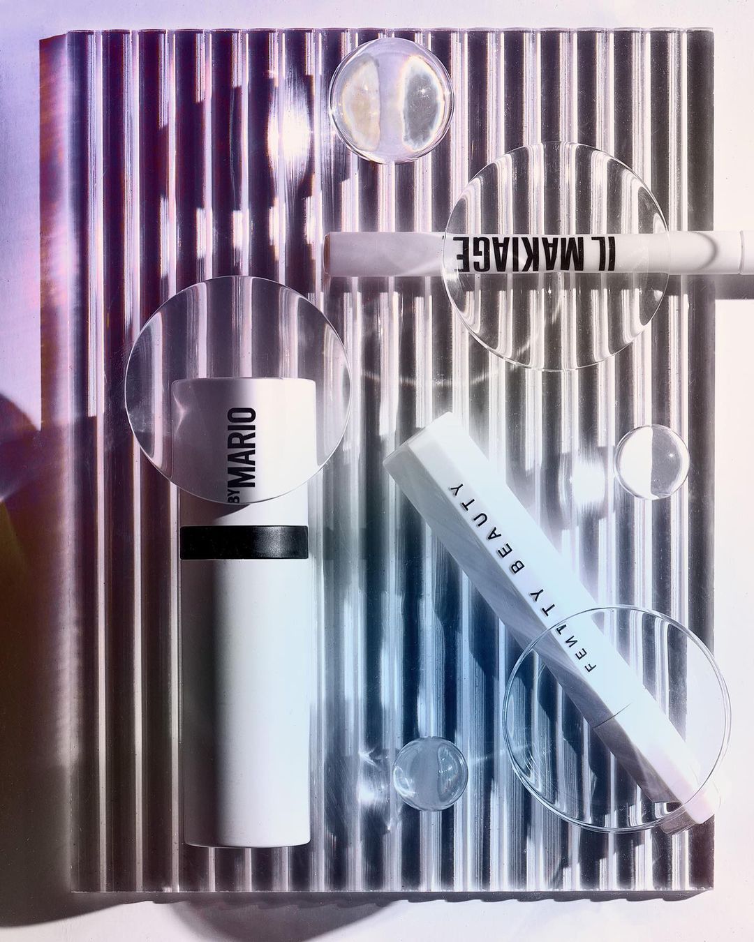 Promotional still life of beauty and makeup products from Fenty Beauty, Il Makiage, and Makeup by Mario on reflective surface.