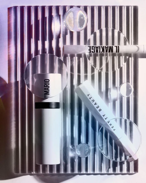 Promotional still life of beauty and makeup products from Fenty Beauty, Il Makiage, and Makeup by Mario on reflective surface.