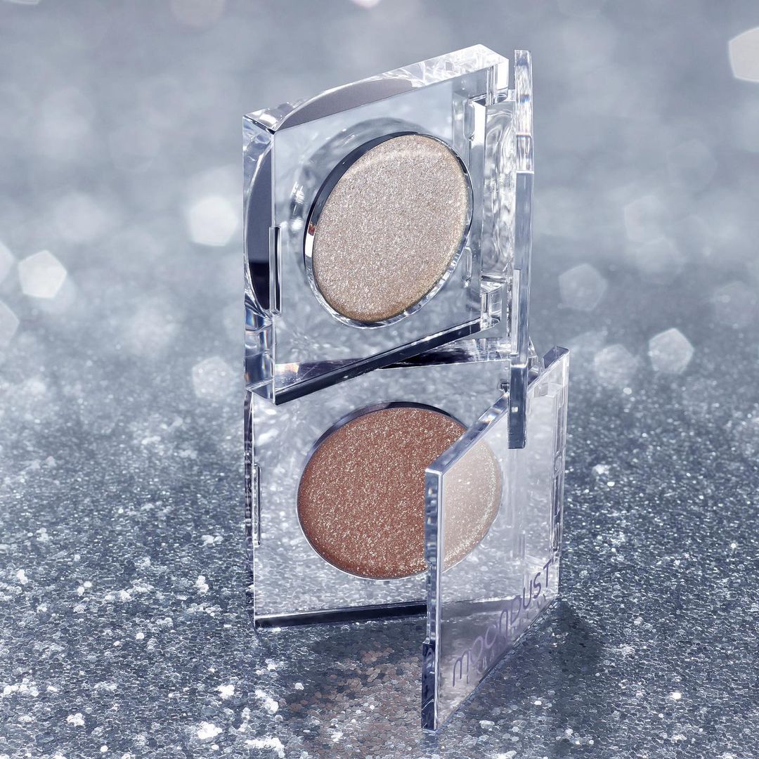 Macro shot of two stacked clear cosmetic containers with golden and bronze eyeshadow, against a sparkling silver background.