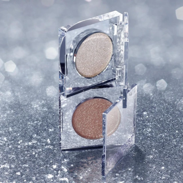 Macro shot of two stacked clear cosmetic containers with golden and bronze eyeshadow, against a sparkling silver background.