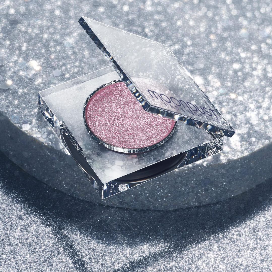 Overhead studio shot of a single, circular, shimmery pink eyeshadow in a mirrored compact, set against a sparkling silver background.