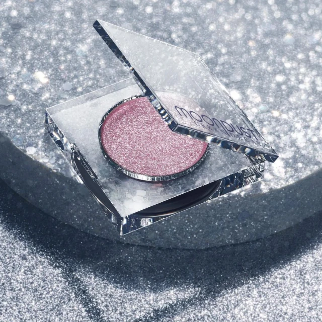 Overhead studio shot of a single, circular, shimmery pink eyeshadow in a mirrored compact, set against a sparkling silver background.
