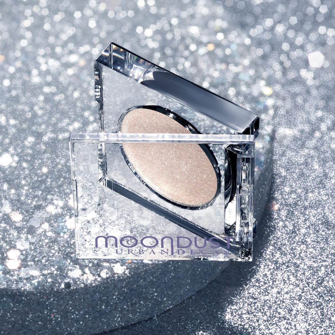 Urban Decay Moondust eyeshadow palette in a clear, square container, showcased against a sparkly silver backdrop, creating a glittery cosmetic still life.