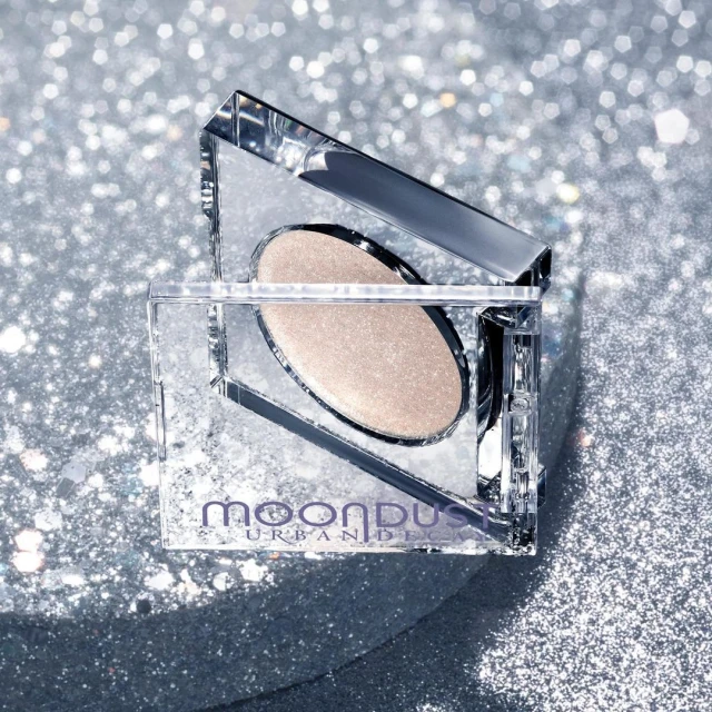 Urban Decay Moondust eyeshadow palette in a clear, square container, showcased against a sparkly silver backdrop, creating a glittery cosmetic still life.