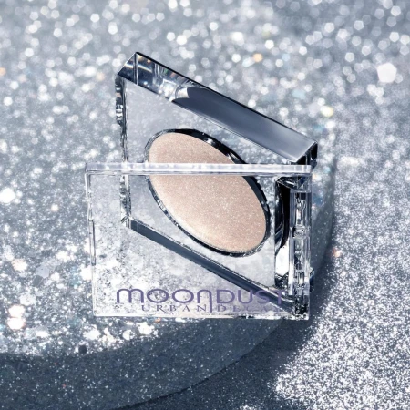 Urban Decay Moondust eyeshadow palette in a clear, square container, showcased against a sparkly silver backdrop, creating a glittery cosmetic still life.