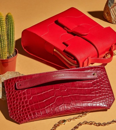Red leather handbags on tan backdrop with cactus. One bag is structured, one is a clutch. Gold chain detail is visible.
