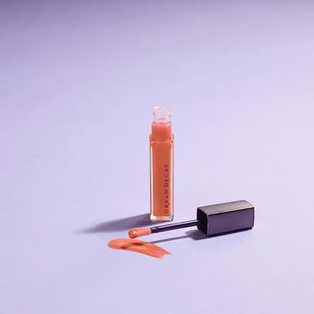 Urban Decay lip gloss tube with applicator and small swatch on a pale lavender surface in a studio shot. Beauty, cosmetics, makeup.