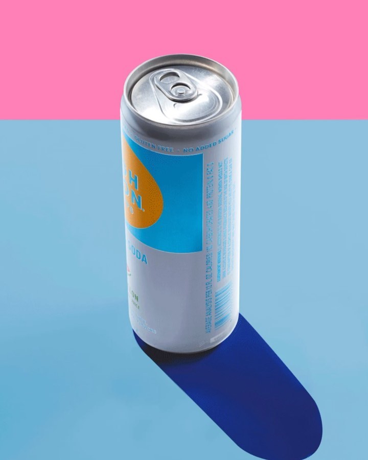 A canned beverage sits on a blue surface with a pink background, creating a vibrant contrast and highlighting the product's design.