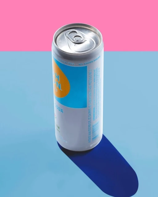 A canned beverage sits on a blue surface with a pink background, creating a vibrant contrast and highlighting the product's design.