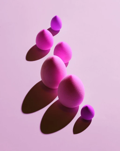 Elevated view of pink makeup sponges in varying sizes arranged diagonally against a light pink backdrop with shadows. Studio shot.