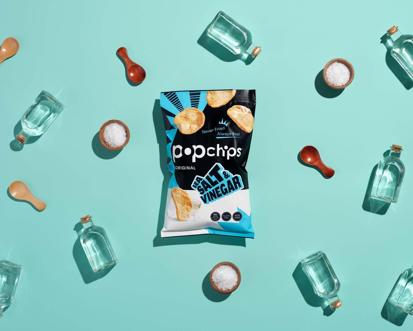 Popchips bag of sea salt and vinegar chips surrounded by wooden spoons, small dishes of salt, and small clear glass bottles on blue surface.