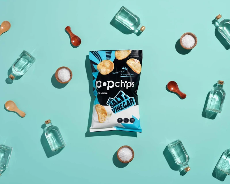 Popchips bag of sea salt and vinegar chips surrounded by wooden spoons, small dishes of salt, and small clear glass bottles on blue surface.