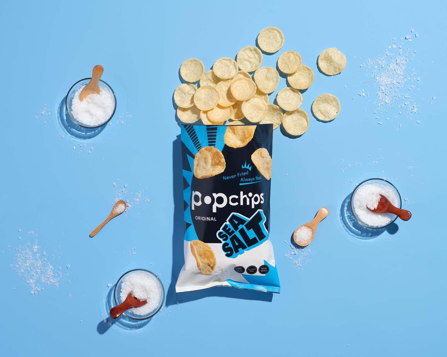 Popchips bag surrounded by potato chips, salt bowls, and wooden spoons on a light blue background, creating a food photography scene.