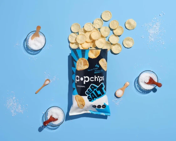 Popchips bag surrounded by potato chips, salt bowls, and wooden spoons on a light blue background, creating a food photography scene.