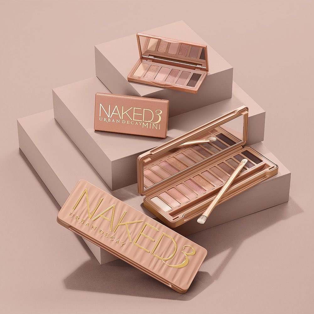 Still life studio shot featuring multiple Naked3 Urban Decay eyeshadow palettes in a warm, neutral color scheme on stepped platforms.